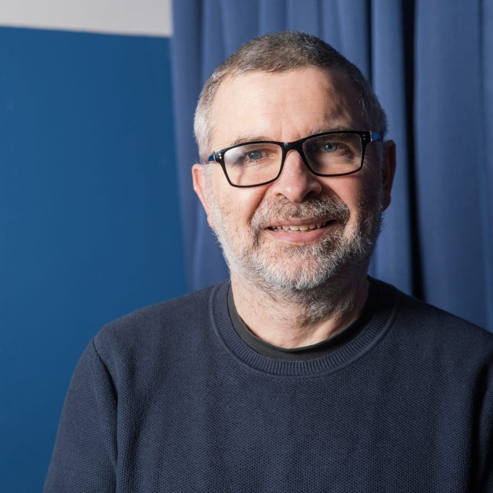 A man with short hair and a beard, wearing glasses and a blue jumper