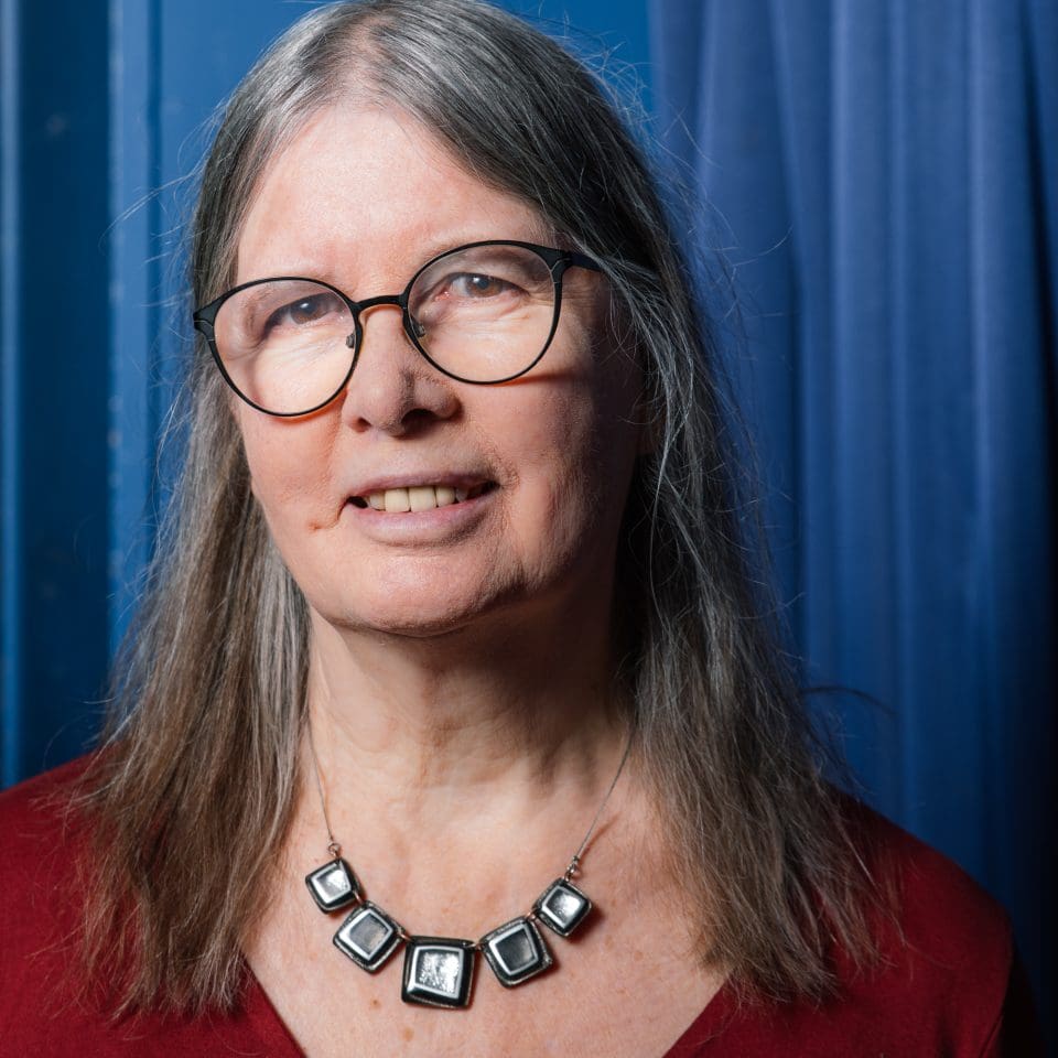 a woman with long grey hair, wearing a red top, glasses and a necklace