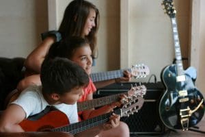 Three children holding guitars look towards a fourth person