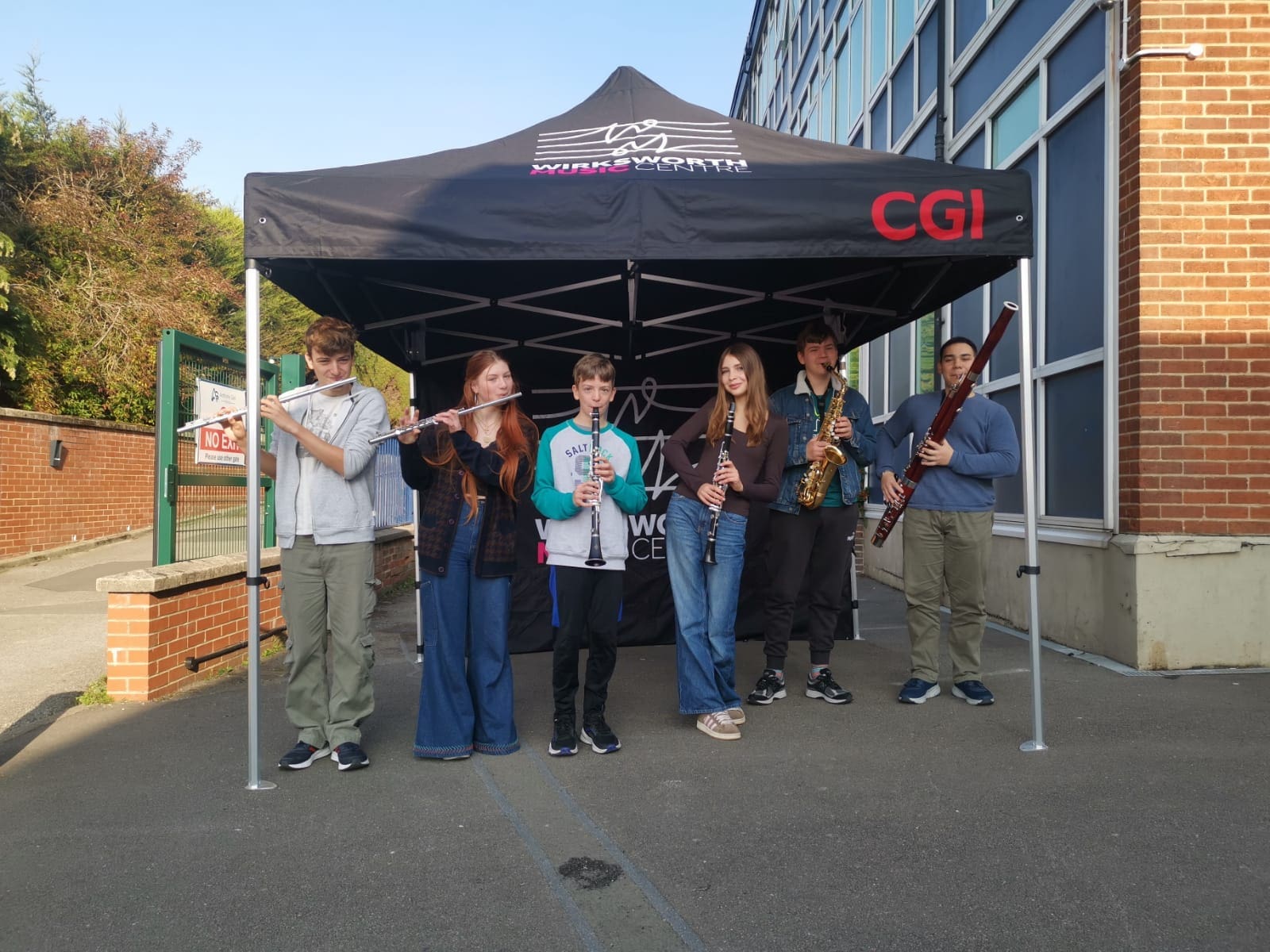 Six young musicians with various wind instruments stand under a black gazebo with the wirksworth music centre and CGI logos on it.